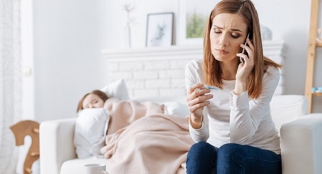 Mum on the phone looking at a thermometer with concern with a child asleep in the background